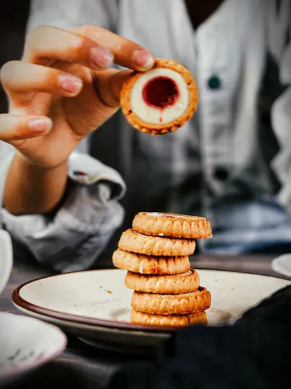 Holding up a single jam-filled cookie to the camera, with a stack on the plate below. This adds a human element and draws the viewer's eye directly to the product's delicious center.
