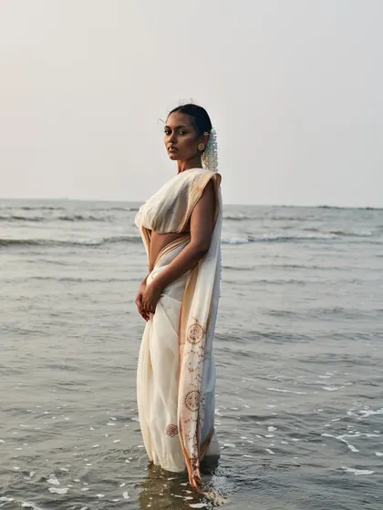 A full-length shot capturing the model standing in the sea, with the saree's fabric flowing around her. The overcast sky and calm water create a serene yet moody backdrop for this editorial photograph.