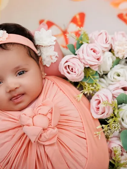 A lovely newborn girl, swaddled in pink, looks at the camera from a bed of flowers. Her alert expression is just priceless.