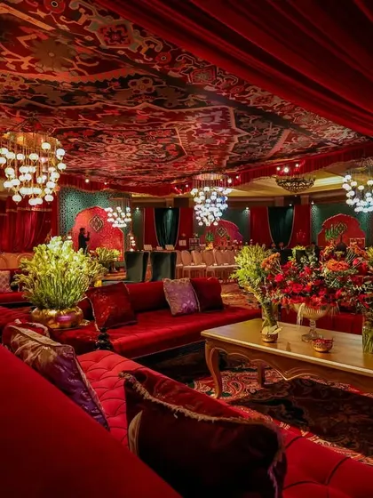 A view across the plush red velvet sofas of the Sufi lounge. The space is filled with floral arrangements and illuminated by clusters of modern chandeliers, blending traditional motifs with contemporary luxury.