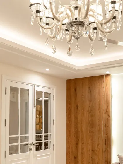 The entrance to this apartment features classic white French doors that open into a living room illuminated by a grand crystal chandelier. The contrast between the white doors and the rich wooden sliding panel adds visual interest.
