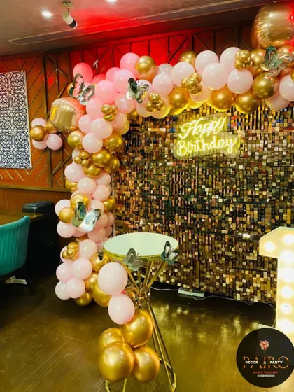 A stylish first birthday setup with a gold shimmer wall. The "Happy Birthday" neon sign, light-up number "1", and cake stand are arranged for a perfect photo corner.