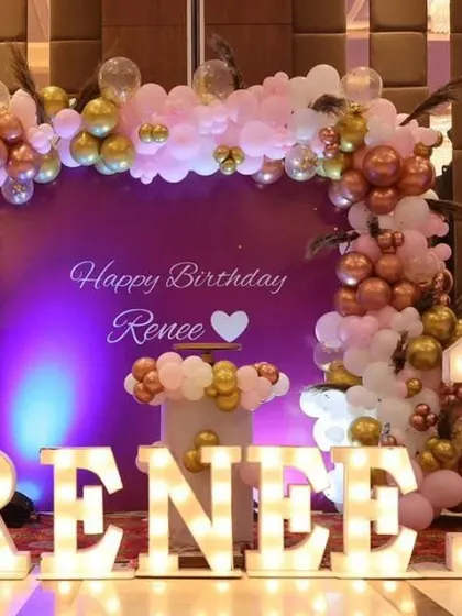 A first birthday setup with large light-up letters spelling the name "RENEE" and a pink and gold balloon backdrop.