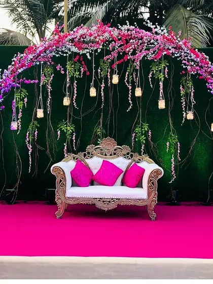 This outdoor stage setup is bursting with color. A deep green hedge wall provides the perfect canvas for a vibrant arch of pink and purple cherry blossoms and hanging lights.