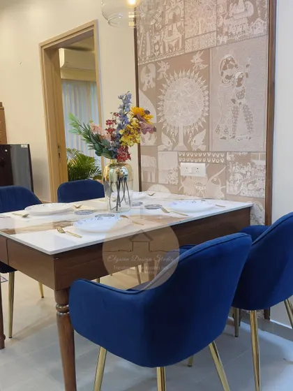 This dining area at Godrej Royale Woods features a beautiful marble-top table, plush blue velvet chairs, and a stunning traditional Indian art wallpaper. The pendant lights add a touch of modern elegance to the space.