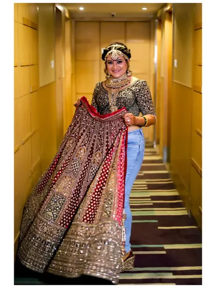 A fun and unconventional shot of the bride holding up her heavy lehenga while wearing jeans underneath. It's a candid, behind-the-scenes moment that shows her playful personality.