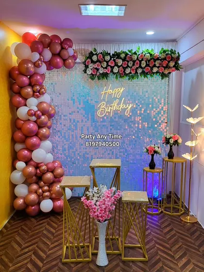 A beautiful party hall decoration featuring a rose gold and white balloon garland against a silver shimmer wall with a floral header.
