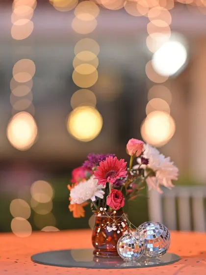 We use lighting and decor to set the mood. This artistic shot captures a colorful floral arrangement and mini disco balls, with the soft bokeh of party lights in the background, hinting at a fun-filled Sangeet night.