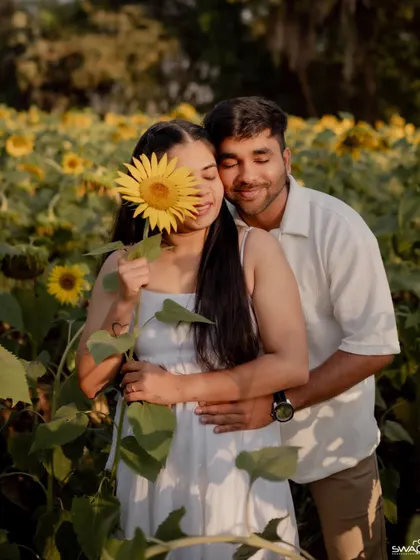 A sweet and intimate pre-wedding portrait where the bride playfully hides her face with a sunflower, while the groom embraces her from behind.