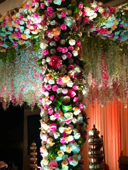 A close-up view of a mandap pillar completely covered in a dense arrangement of pink, peach, and white flowers. This technique creates a lush, textured look and feel, complemented by hanging white floral strings.