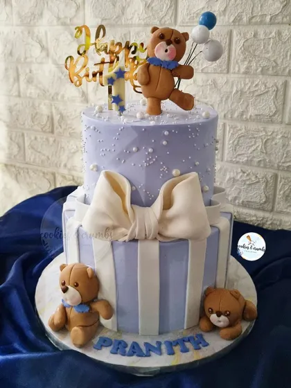An adorable two-tiered teddy bear cake for a first birthday. The design includes a big fondant bow, stripes, and cute bears, creating a classic and sweet look for a little boy.