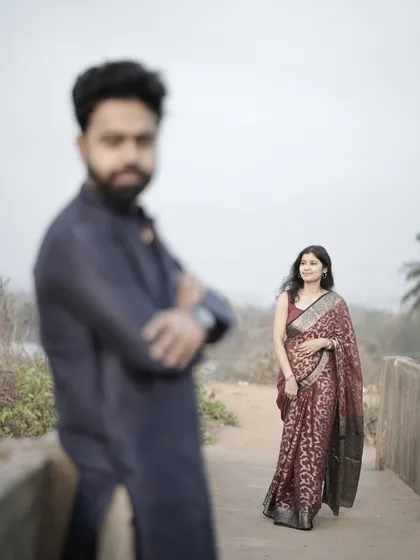 An artistic shot with the groom out of focus in the foreground, drawing the eye to the bride in her beautiful red saree.