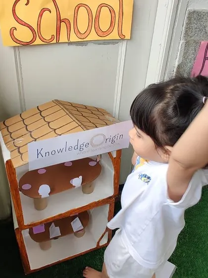 A child points to the miniature "Knowledge Origin" schoolhouse in our role-play setup, showing pride in her community.
