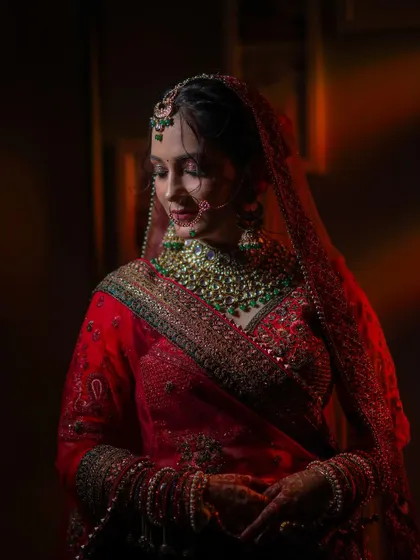 A classic bridal portrait showing the bride in her stunning red lehenga. The soft, dramatic lighting highlights her graceful pose and the richness of her wedding attire.
