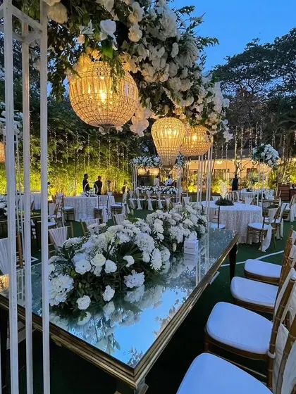 An elegant engagement dinner setup with a white and green theme, featuring long mirrored tables, lush floral runners, and beautiful overhead chandeliers.