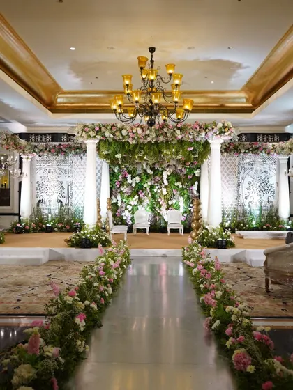 A breathtaking wedding aisle leading to a stage covered in a canopy of white and pink florals. The mirrored floor reflects the floral ceiling, creating a path that feels like walking through a garden in the sky.