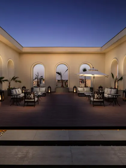 The terrace of a neoclassical residence at dusk. The warm lighting, classical arches, and comfortable seating create an inviting open-air lounge, perfect for serene evenings.