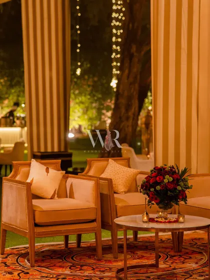 A cozy lounge setup with modern wooden-framed chairs and a Persian-style rug. The space is made intimate with bouquets of red roses and the soft glow of background lights.