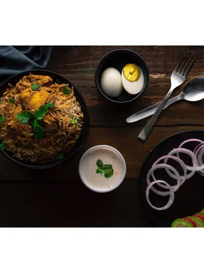A complete biryani meal composition, shot on a dark wooden surface to create a rustic, traditional feel. The arrangement includes side dishes like raita and boiled egg, showing a full serving suggestion.