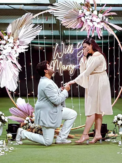 A beautiful moment captured. This modern proposal features a gold ring backdrop decorated with pampas grass and white florals, with delicate fairy lights and a neon sign to complete the romantic, boho-inspired look.