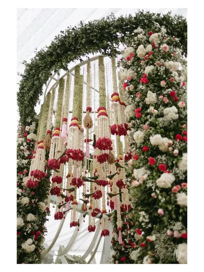 A close-up of the floral archway from a grand wedding ceremony. The dense arrangement of white, pink, and red flowers creates a rich texture and a stunning frame for the mandap.