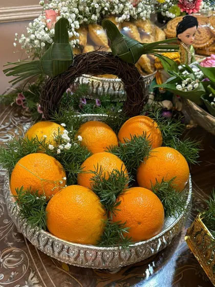 This rustic-themed fruit tray features oranges arranged in a silver platter with a woven twig wreath as a handle. The small, handmade birds made from coconut leaves add a charming and unique touch to the design.