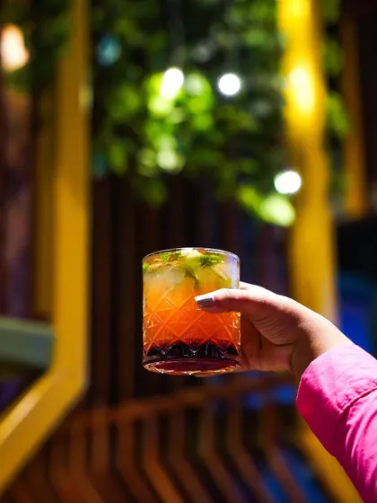 A hand holds a layered orange and red cocktail against a softly blurred background of a bar. This simple, elegant shot focuses on the drink and creates a relaxed, social atmosphere.