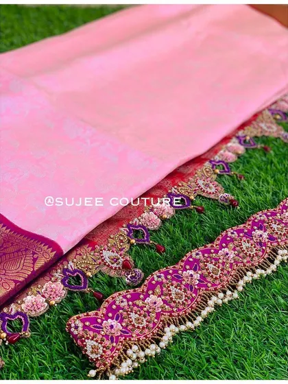 A beautiful flat-lay showcasing the components of a custom half saree. It includes a baby pink saree, a heavily embroidered purple vaddanam, and matching tassels with heart-shaped details.