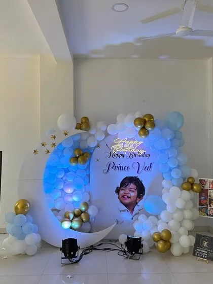 This moon-themed birthday decoration includes a personalized backdrop with the child's photo. The setup is complemented by a milestone photo board, flower vases, and a blue and white balloon garland with gold accents.