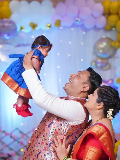 A joyful family moment from a naming ceremony. The proud father lifts his baby high in the air as both parents look on with love and happiness.