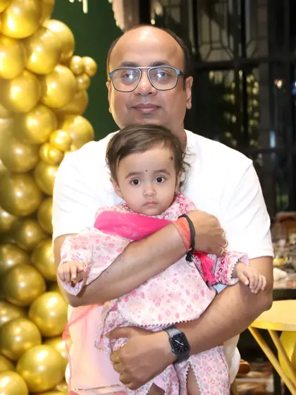 A father holds his daughter in front of a wall of golden balloons at her party.