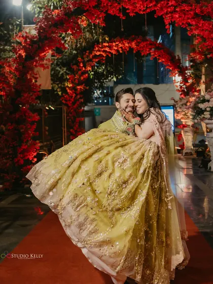 The groom lifts his bride in his arms under a stunning red floral archway, a romantic and fun moment from their Sangeet.
