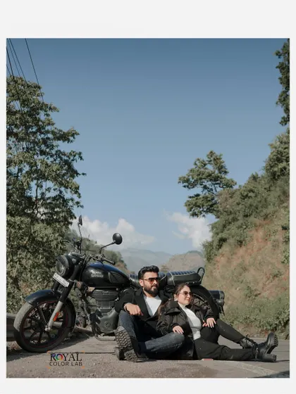 A wide-angle shot of a couple sitting with their motorcycle against a clear blue sky and lush green hills, capturing the freedom of the open road.