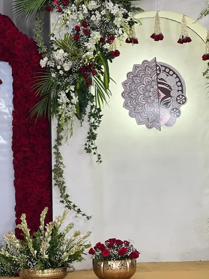 A corner detail from the "Mitakshi" naming ceremony. The Maa Durga art piece is illuminated, flanked by lush greenery and red roses, creating a sacred and visually stunning focal point.