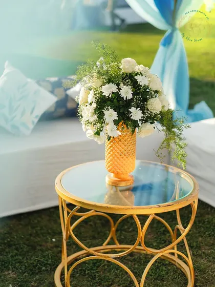 A detail shot of a side table at the Mehendi Sangeet. A gold vase with white flowers adds a touch of glamour to the outdoor setting.