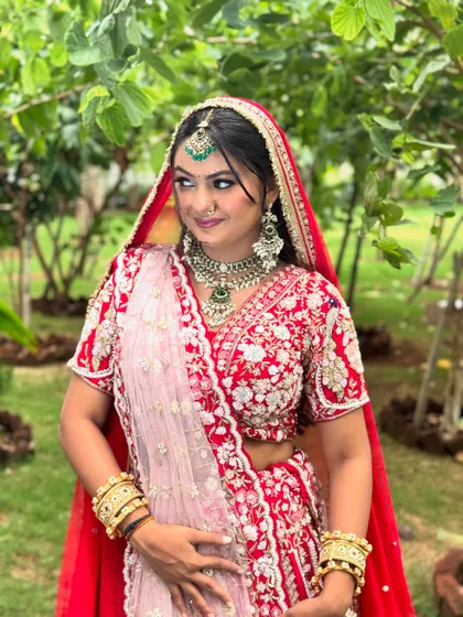 A beautiful portrait of a bride in her red wedding attire, surrounded by lush greenery. The makeup is designed to look fresh and vibrant in natural daylight.