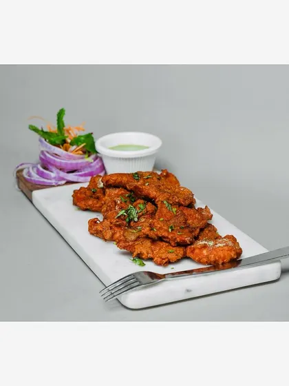 A platter of fried chicken appetizers with a side of mint chutney and salad, presented on a white marble board. The clean, light grey background ensures the food is the hero.