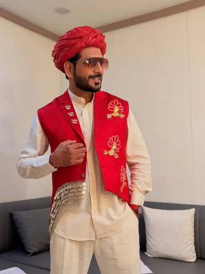A classic Rajasthani look with a red bandhgala jacket, pagdi, and a traditional kurta. The coin detailing on the jacket adds a unique, festive touch.