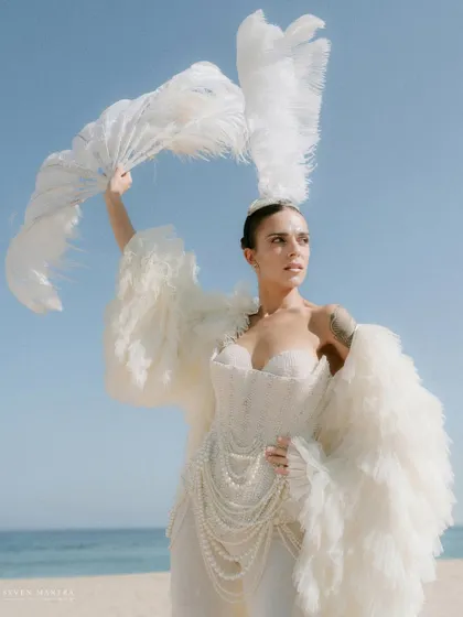 A goddess of the sea. This dramatic look, styled for a beach carnival, features a structured white corset, a flowing feather coat, and a dramatic feather fan, creating an unforgettable, high-fashion moment against the blue sky.