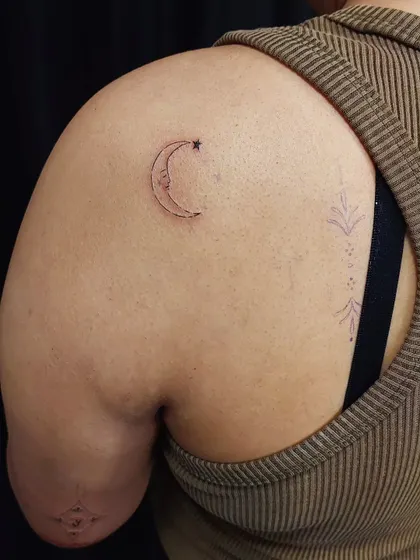 A whimsical crescent moon and star tattoo on the shoulder. The fine-line work gives it a delicate, hand-drawn feel.
