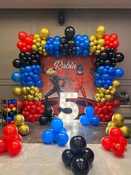 A Miraculous Ladybug themed 5th birthday party. The decor uses the signature red, blue, and black colors, with a character backdrop and a light-up marquee number.