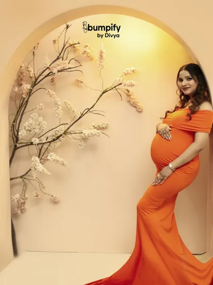 A vibrant and modern studio portrait. The mom-to-be looks stunning in a bright orange form-fitting gown, posed against our minimalist floral arch set.