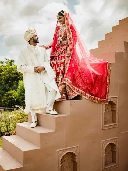 A creative pose on the steps of the venue. The bride stands above the groom, her red lehenga flowing down, creating a visually interesting and dynamic portrait.
