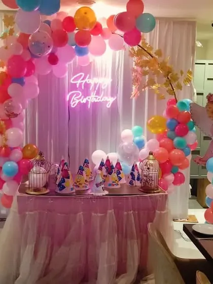 An adorable angel-themed birthday party. The decor features a round balloon arch in pastel colors, a 'Happy Birthday' neon sign, and cute angel cutouts on balloon pillars next to the cake table.