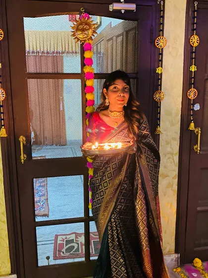 Holding a plate of diyas for Diwali, dressed in a traditional dark silk saree with a contrasting red blouse. This is a classic festive look that is both elegant and timeless.