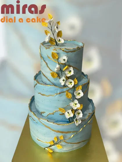 A breathtaking three-tier wedding cake with a blue marble effect and gold veining. The cake is adorned with delicate branches of white sugar flowers, creating a serene and elegant look.