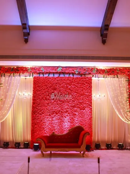 A bold and beautiful reception stage featuring a dense red floral wall. Paired with a classic red chaise lounge and soft white drapes, this design makes a dramatic and passionate statement.
