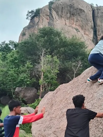 A young climber wearing a helmet carefully plans her next move on a boulder, with fellow students ready to spot her.