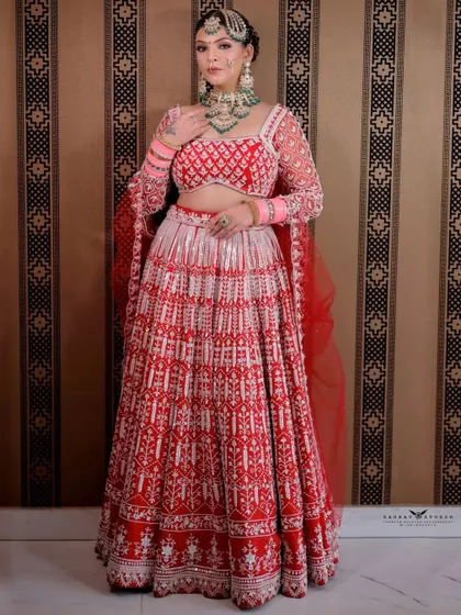 A full-length shot of the bride in a stunning red and white embroidered lehenga. The simple background makes the intricate details of the outfit stand out.