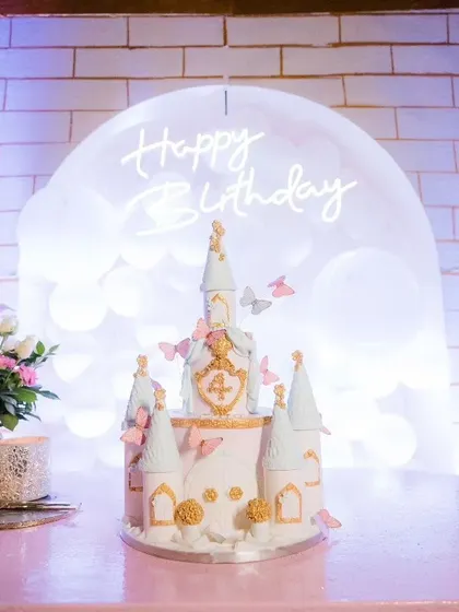 A fairytale castle cake is the ultimate showstopper. This one features delicate butterfly details and sits in front of a custom 'Happy Birthday' neon sign, creating a perfect photo moment.